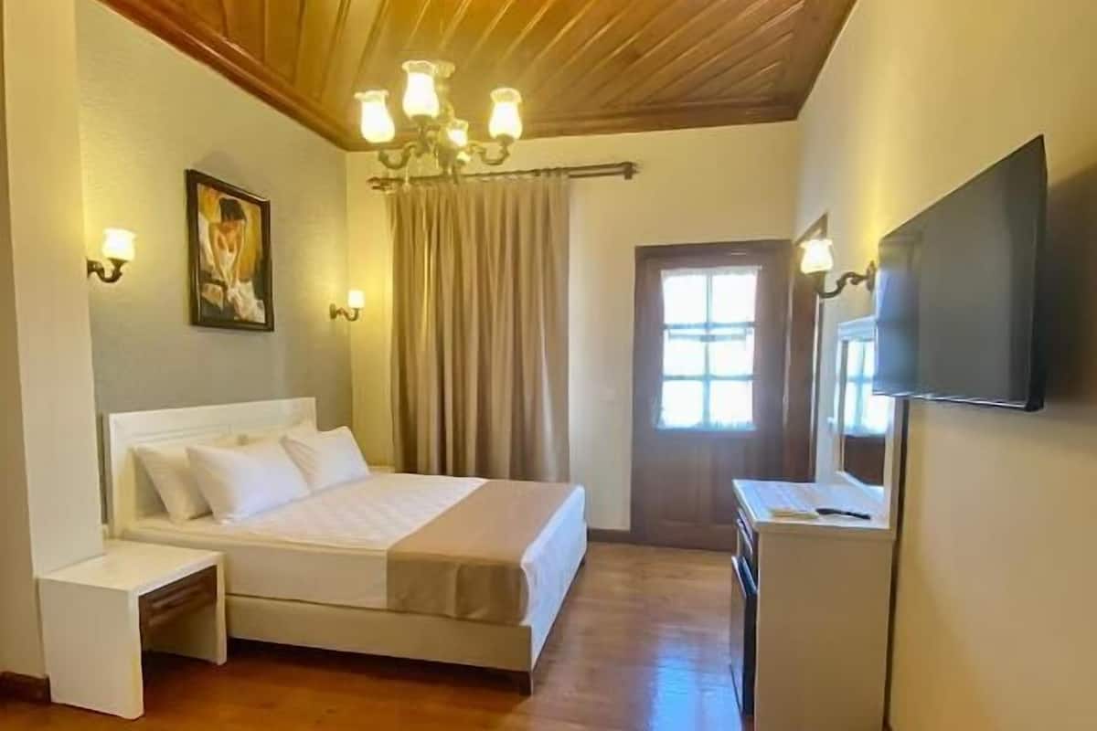 A bedroom with a wooden ceiling, a bed, bedside tables, a desk, a TV, and a window with curtains.