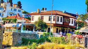 A coastal town with a prominent building featuring a tiled roof and white walls, adjacent to a stone wall and a restaurant with outdoor seating. - Ulivo Hotel (Antalya)