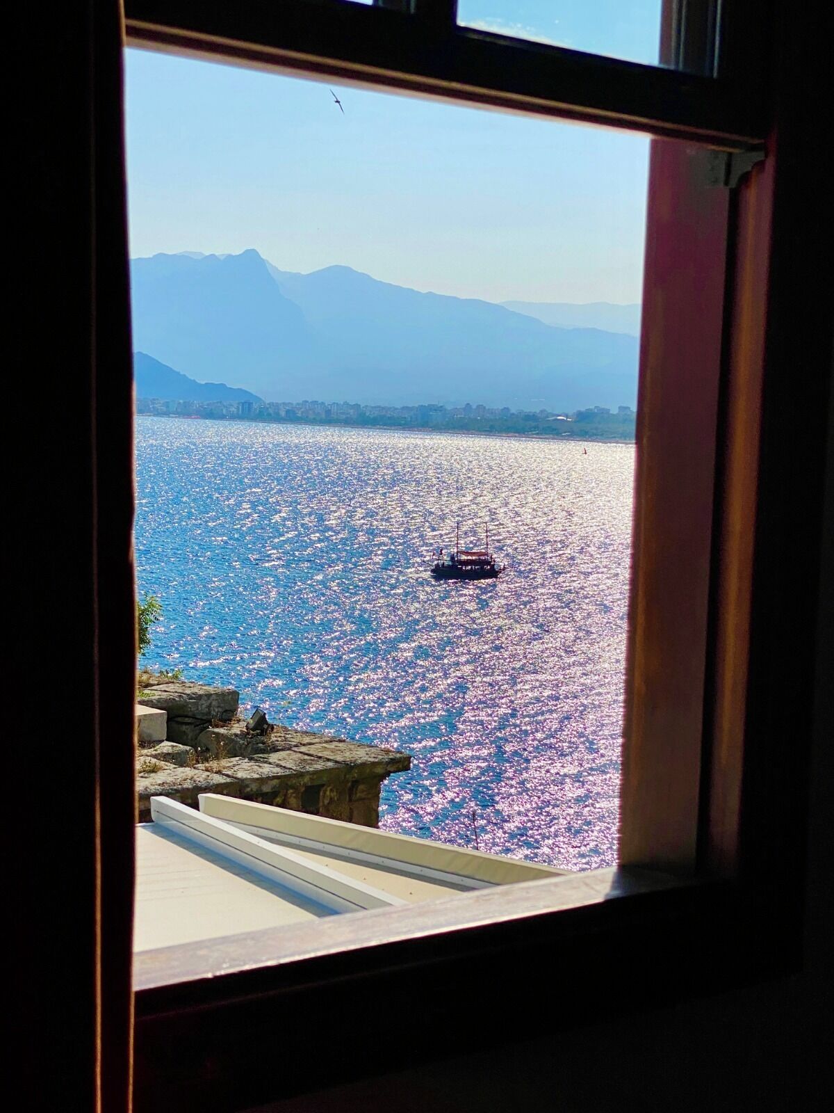 A view through a window of a coastal scene with a boat on the water, mountains in the background, and a rocky shoreline.