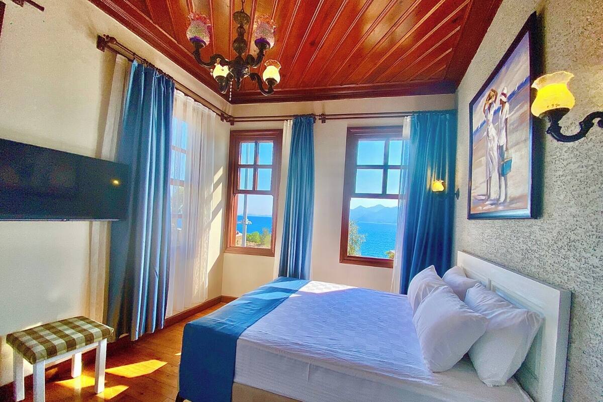 A bedroom with a bed, a bench, two windows with curtains, a chandelier, and a painting on the wall.