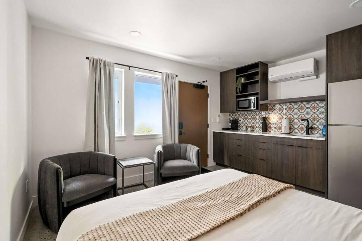 A hotel room with a bed, two chairs, a small table, a kitchenette with a microwave, and an air conditioning unit.