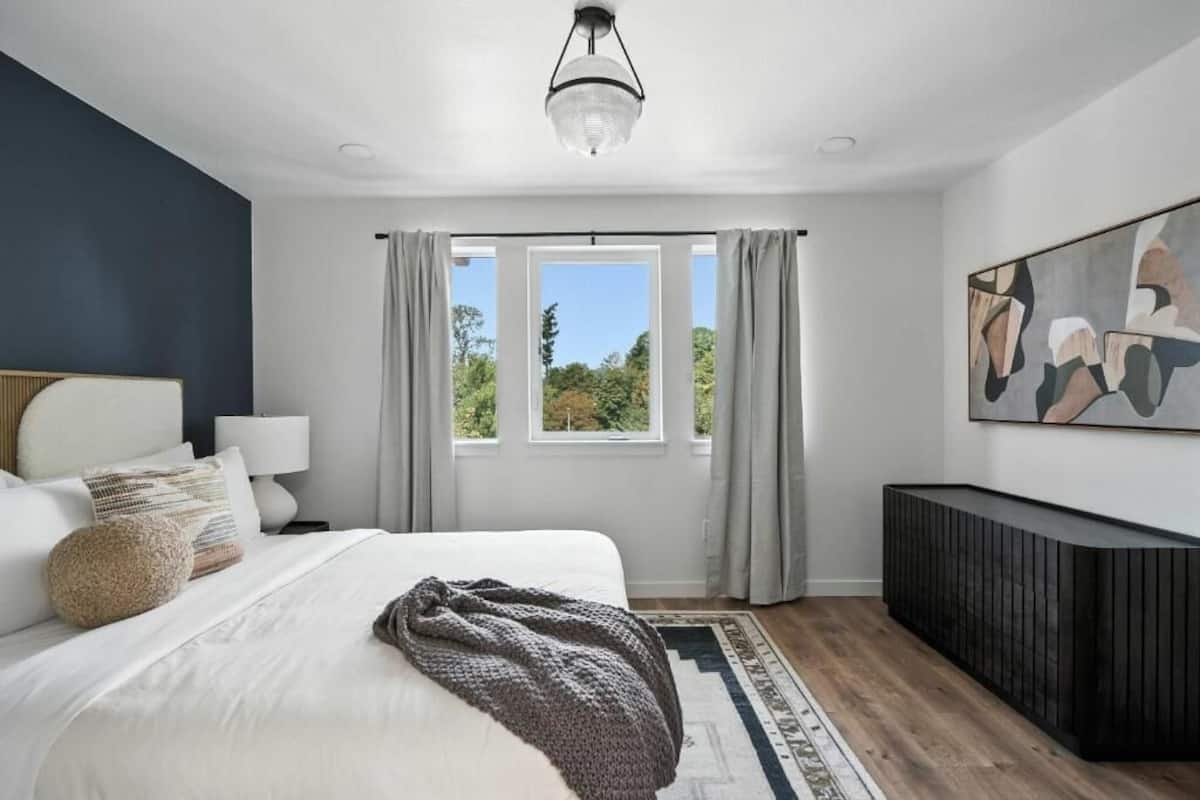 A bedroom with a bed, a bedside table, a lamp, a window with curtains, a painting on the wall, and a black console.