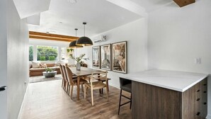Una cocina moderna con encimera blanca, mesa de comedor de madera y lámparas colgantes.