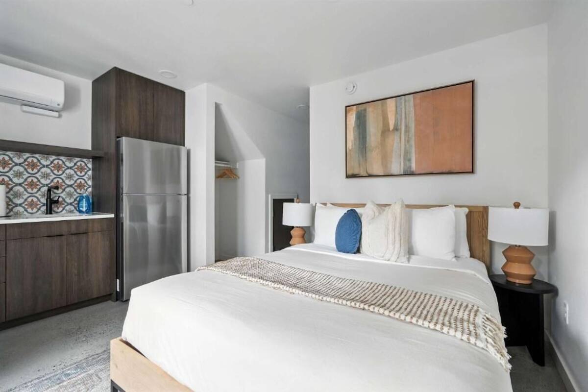 A modern hotel room with a bed, a kitchen area, and a large abstract painting on the wall.