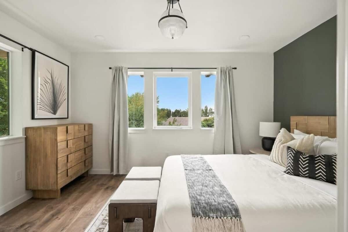 A bedroom with a large bed, a wooden dresser, a nightstand with a lamp, and a window with curtains.