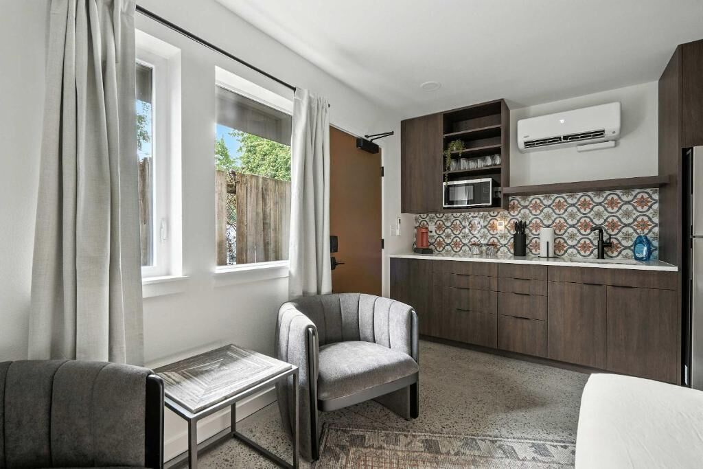 A compact kitchen with a built-in oven, a microwave, and a sink. There is a window with curtains, a small table, and a chair.