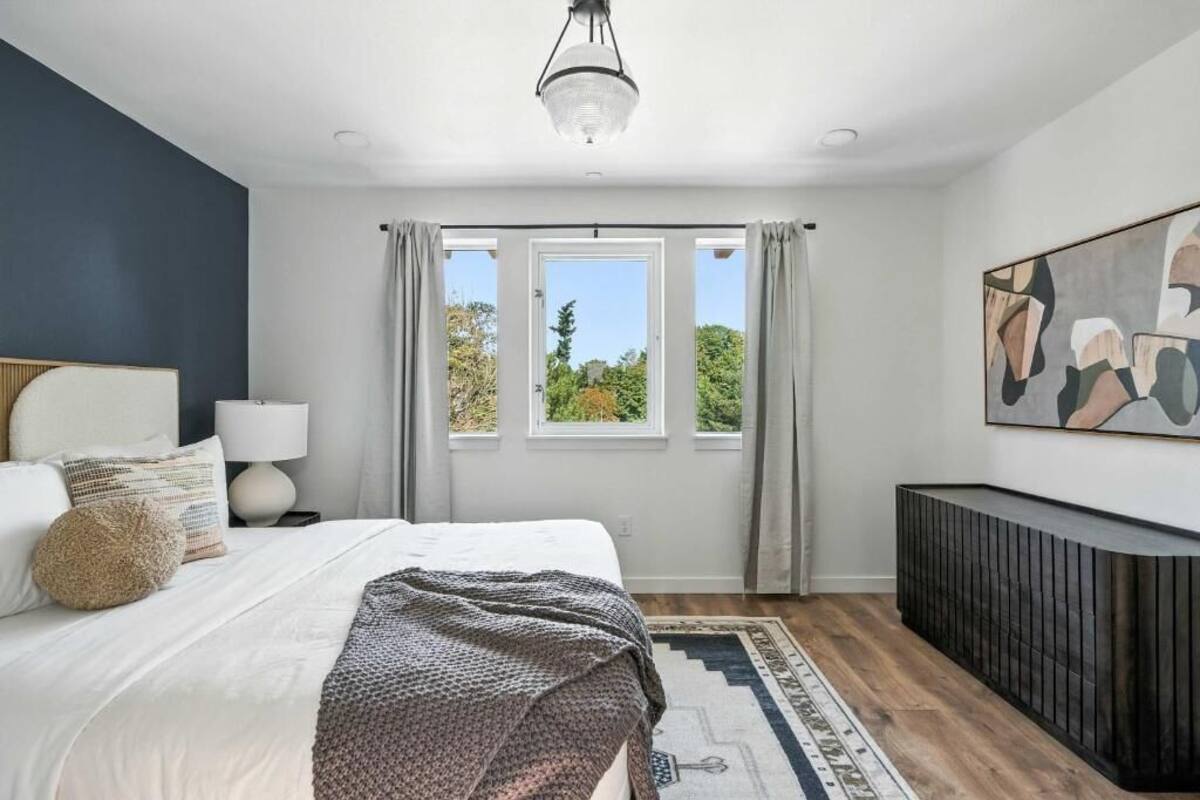 A bedroom with a bed, a bedside table, a lamp, a window with curtains, a painting on the wall, and a console table.