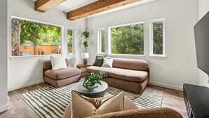 Una sala de estar moderna con un conjunto de sofá, una mesa de centro con una planta y ventanas amplias que ofrecen vistas a espacios verdes.