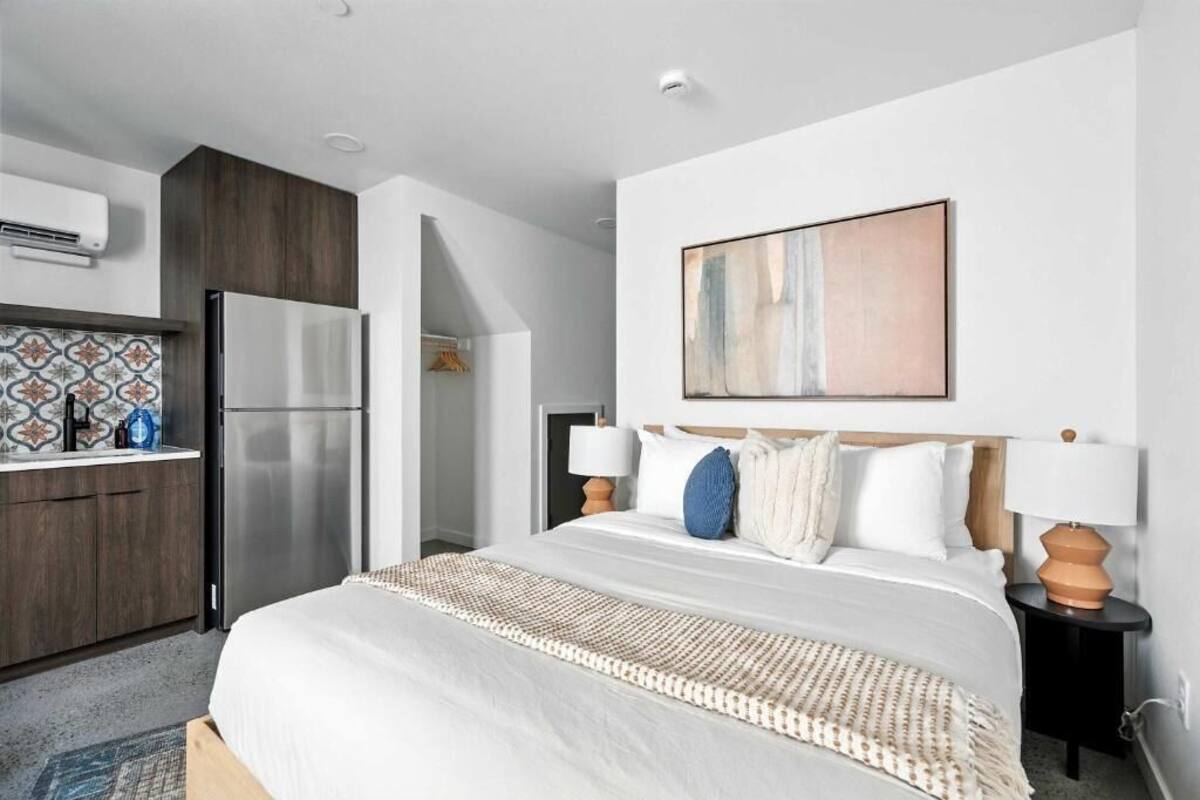 A hotel room with a bed, bedside tables, a wall-mounted artwork, a kitchen area with a refrigerator, and a patterned backsplash.