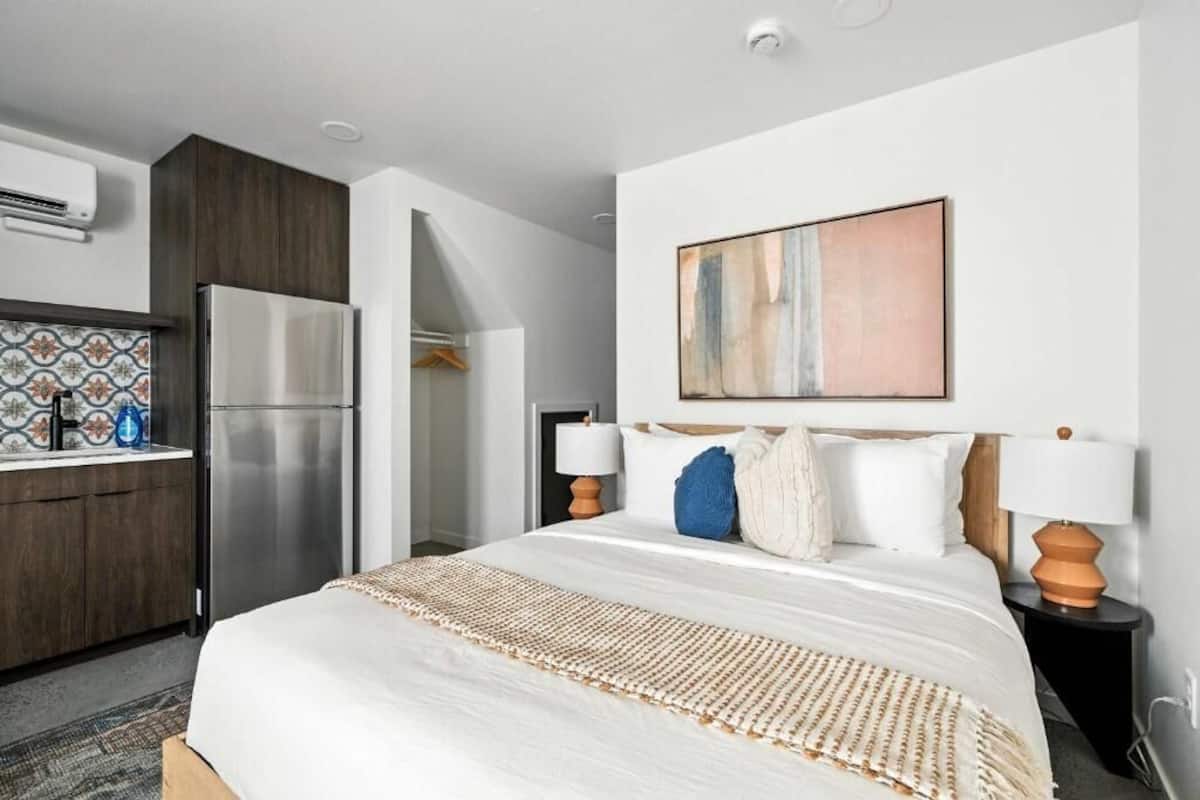 A hotel room with a bed, a nightstand, a lamp, a refrigerator, a kitchen area, and a wall-mounted artwork.