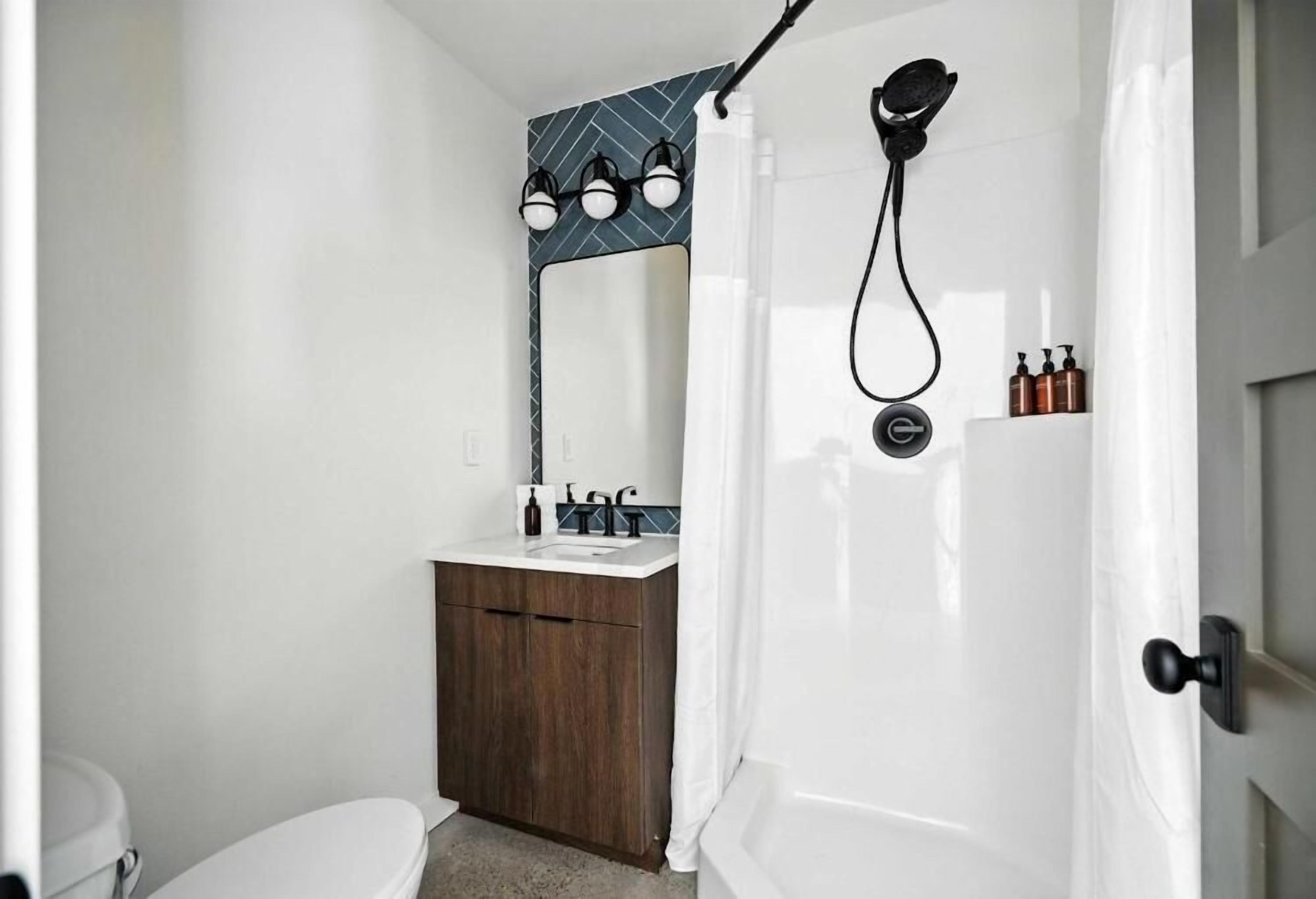 Un baño con inodoro blanco, tocador de madera, espejo, ducha con cabezal de ducha manual y perchero para toallas.