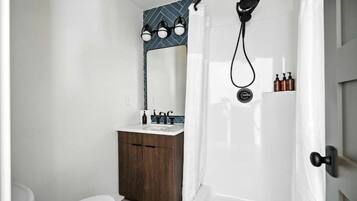 Un baño con inodoro blanco, tocador de madera, espejo, ducha con cabezal de ducha manual y perchero para toallas.