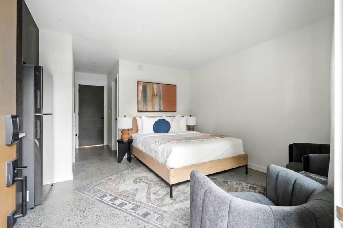 A modern hotel room with a bed, a seating area, and a kitchenette.