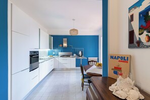 A modern kitchen with white cabinets, a dining area with a wooden table, and a blue accent wall. - Linda Blue Bay, Sorrento Coast (Piano di Sorrento)