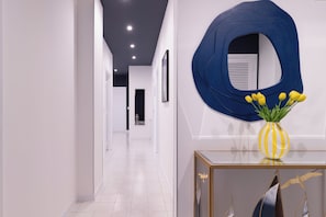 Un couloir moderne aux murs blancs, agrémenté d’une œuvre d’art abstraite bleue et d’un vase jaune contenant des fleurs jaunes, posé sur une console.