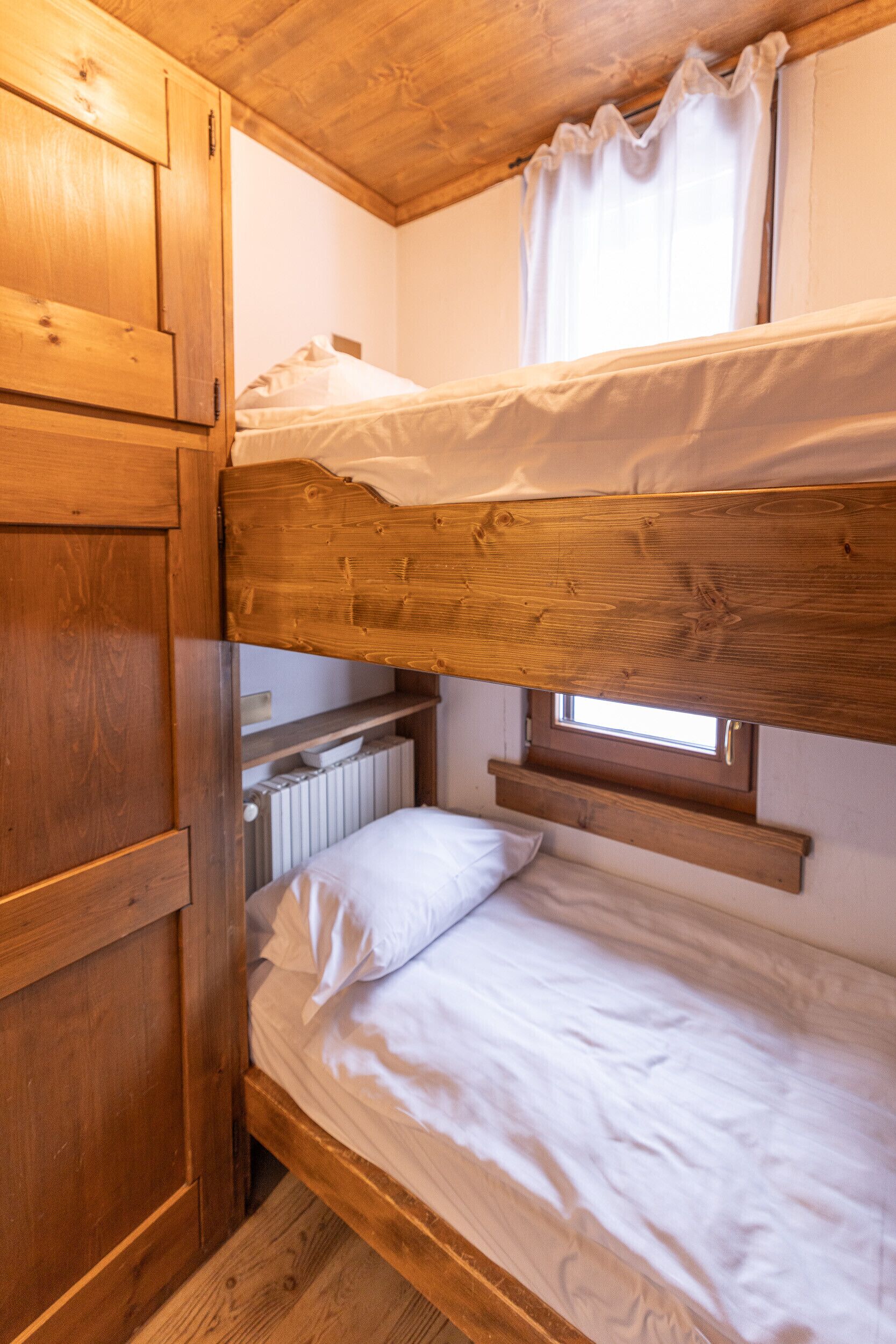 A bunk bed with wooden headboard and footboard, a window with white curtains, and a radiator below the window.