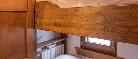 A bunk bed with wooden headboard and footboard, a window with white curtains, and a radiator below the window.