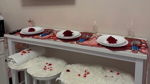 Una stanza con una TV montata a parete, un tavolo apparecchiato con una rosa rossa e due sgabelli bianchi e soffici.