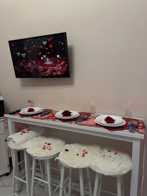 A room with a TV mounted on the wall, a table set with a red rose, and two white furry stools. - House in the ancient heart (Napoli)