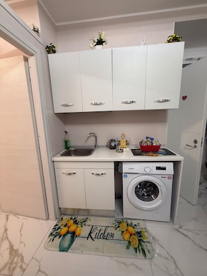 Une cuisine compacte avec des meubles blancs, un lave-linge encastré, un évier et un tapis décoratif sur lequel est inscrit le mot « Cuisine ».