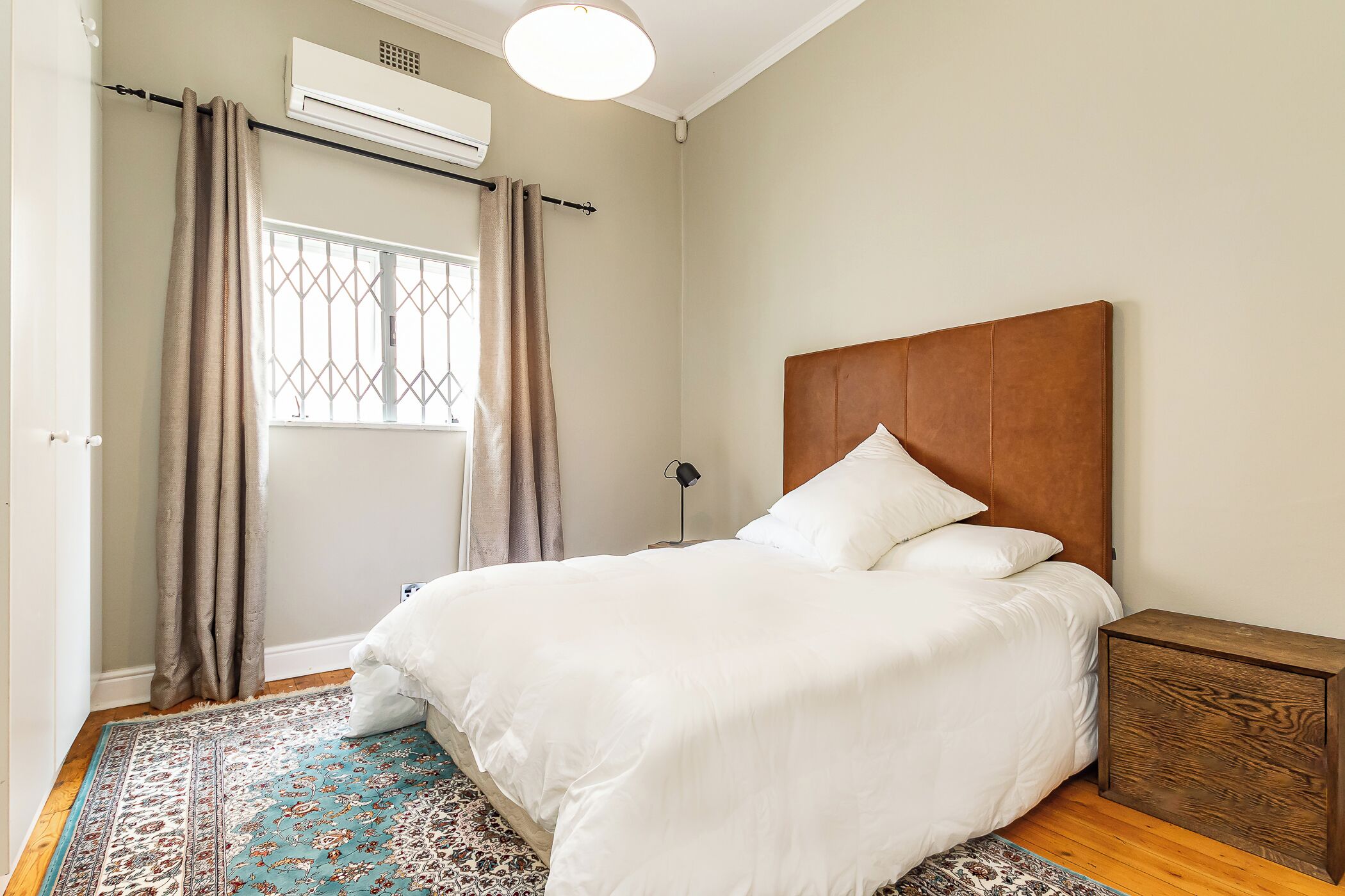 A bedroom with a bed, curtains, an air conditioning unit, a bedside table, and a patterned rug.