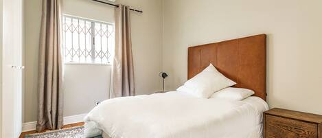A bedroom with a bed, curtains, an air conditioning unit, a bedside table, and a patterned rug.