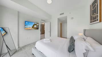 A modern hotel room with a large bed, a flat-screen TV mounted on the wall, and a view of the beach through the window.