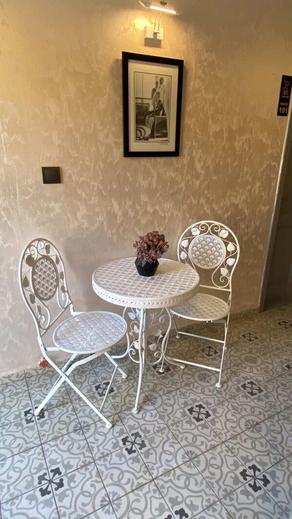 Uma pequena mesa de metal branca e duas cadeiras a combinar, com um vaso de flores secas sobre a mesa, dispostas em frente a uma parede com textura e uma moldura acima.