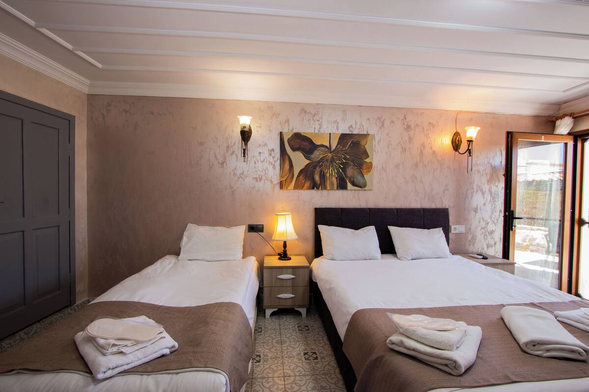 A hotel room with two beds, a nightstand with a lamp, a painting on the wall, and a view through the door.