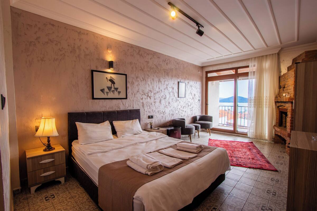 A hotel room with a large bed, a nightstand with a lamp, a chair, a fireplace, and a view of the outdoors.