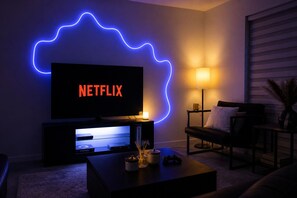 Ein Wohnzimmer mit Fernseher, auf dem 'NETFLIX' angezeigt wird, einer Lampe, einem Sofa und einem Couchtisch mit Dekorationselementen.