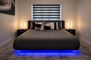 Ein modernes Schlafzimmer mit einem großen Bett, zwei Nachttischen, einem Fenster mit Jalousien und einem blau beleuchteten Boden.