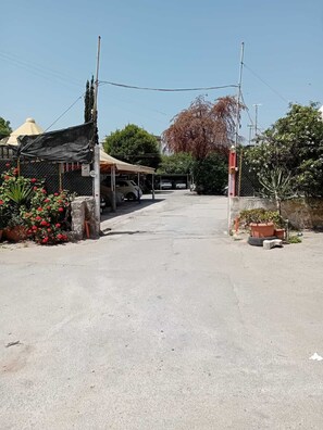 Un camino pavimentado que conduce a un edificio con un porche cubierto, posiblemente un garaje o taller, y una zona vallada con plantas en macetas.