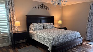 A bedroom with a wooden floor, a bed with a patterned bedspread, two bedside lamps, a chandelier, and a decorative wall piece. - South Alabama Country Home With A Swimming Pool (Fairhope)