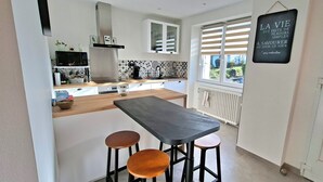 A modern kitchen with a central island, bar stools, a black countertop, and a window with blinds. - Renovated country house close to the ocean and hiking trails. (Névez)