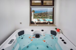 Un bain à remous doté d’un panneau de commande, une fenêtre donnant sur une piscine et un paysage, et un objet décoratif posé sur le rebord de la fenêtre.