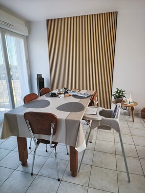 A dining area with a table set for four, chairs, a potted plant, and a small table with a plant and a candle. - Ty Macareux: Peace, Comfort & Terrace—15 Minutes from the Beach/GR34 (Lannion)