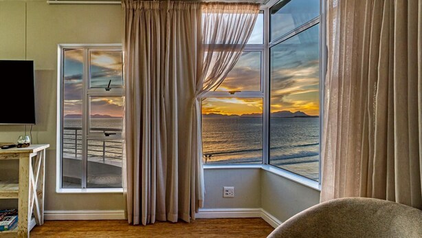 A room with a large window overlooking a beach, a television, and a wooden floor. - 75 Cap D'Azur (Cape Town)
