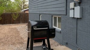 Una parrilla negra con ruedas, ubicada en un patio de concreto al lado de una casa con revestimiento gris y una pared de ladrillo.