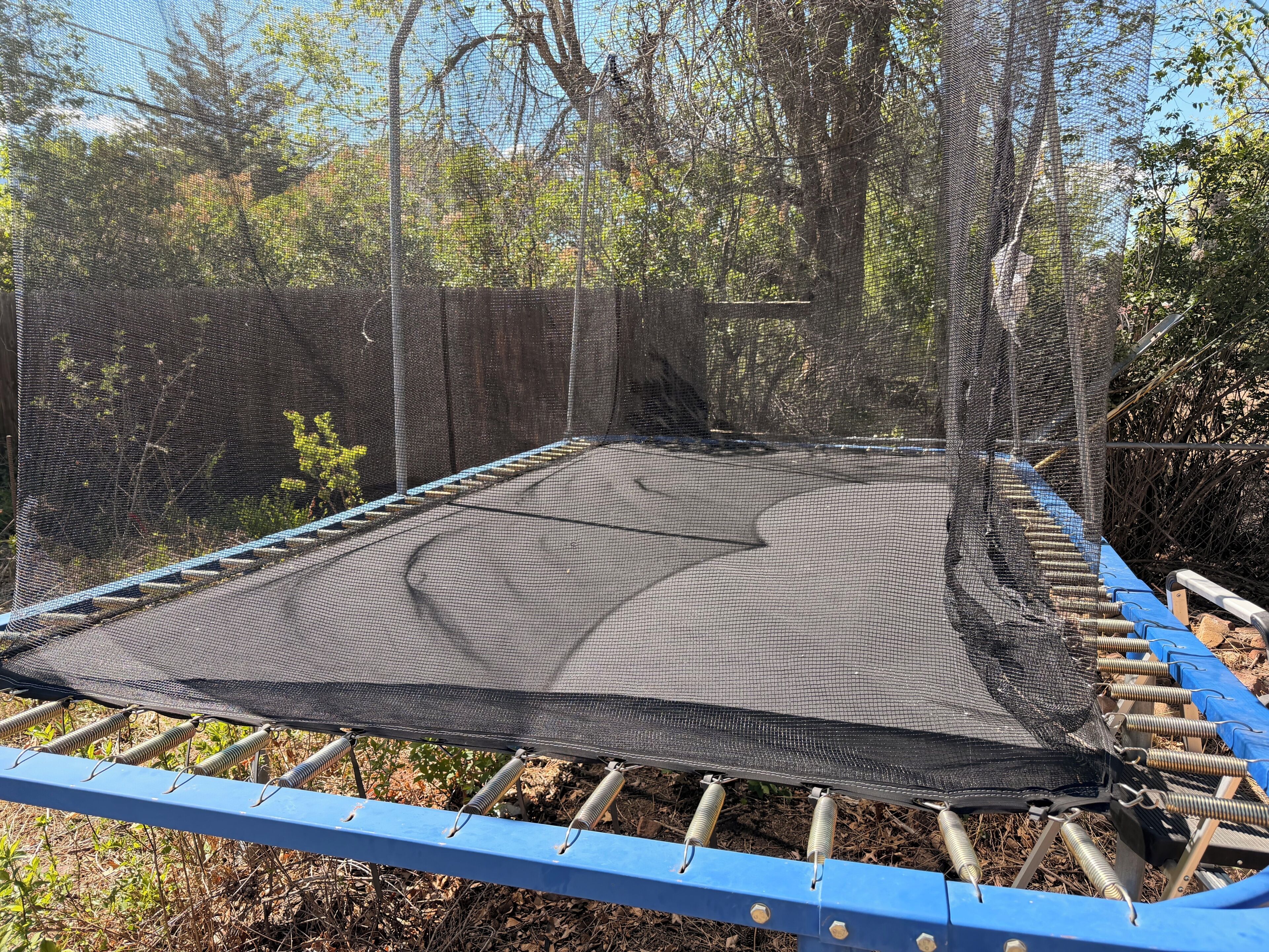 Trampolí amb xarxa de seguretat, envoltat d'arbres i una estructura metàl·lica blava.