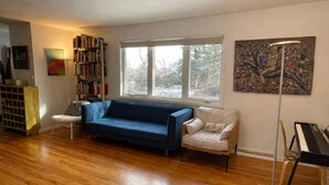 Una sala de estar con un sofá azul, un sillón blanco, una estantería, un piano y un ventanal grande.