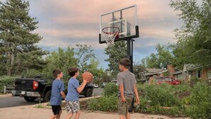 Cancha de baloncesto al aire libre en una zona residencial, con un aro, árboles y vehículos estacionados.