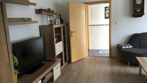 Una sala moderna con televisor de pantalla plana, estanterías de madera, un sofá y una puerta que da a otra habitación.