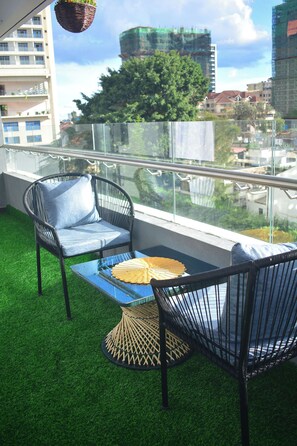 Un balcon avec un revêtement de sol en gazon synthétique, deux fauteuils en osier avec des coussins, une table en verre avec un set de table en fibres tressées, et une vue sur des immeubles et des arbres.