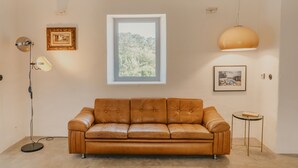 A room with a brown leather sofa, a floor lamp, a framed picture, a pendant light, and a window with a view of trees. - Exclusive Villa Serra de Baixo - Porto Covo (Setúbal)