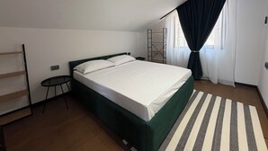 Een enkel bed met een groen hoofdeinde in een kamer met een schuine plafond, een klein nachtkastje, een zwarte stoel en een gestreept vloerkleed.