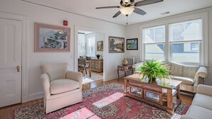Ein Wohnzimmer mit Deckenventilator, einer Couch, einem Couchtisch und einem großen Fenster mit Jalousien.