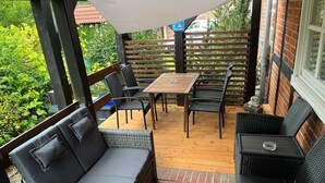 A covered patio with wicker furniture, a wooden table, and a brick wall.
