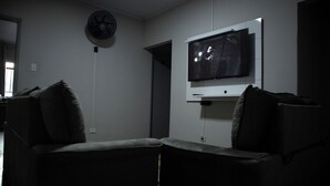 A dimly lit room with a television mounted on the wall, a standing fan, and a couch with pillows. - Hostel Estação da XV (Telêmaco Borba)