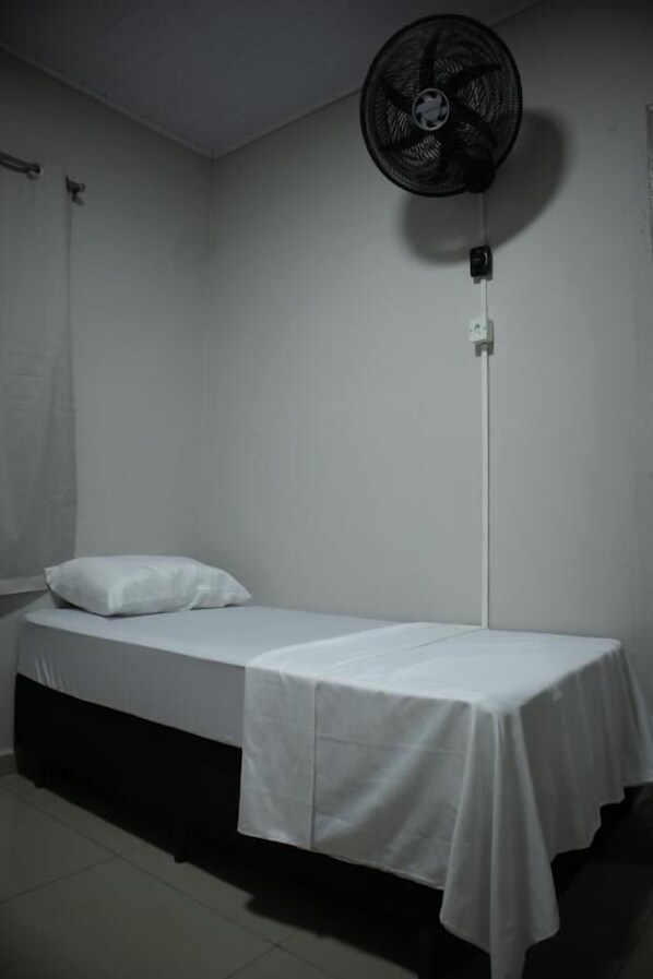 Una cama individual con una colcha blanca y almohada, un ventilador de pie y una pared blanca.